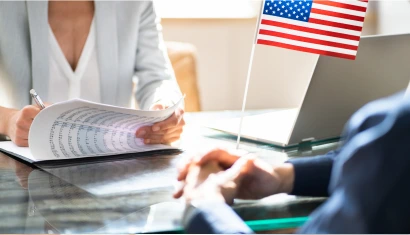 A woman in a light grey blazer signing a document at a glossy table, an American flag beside her laptop